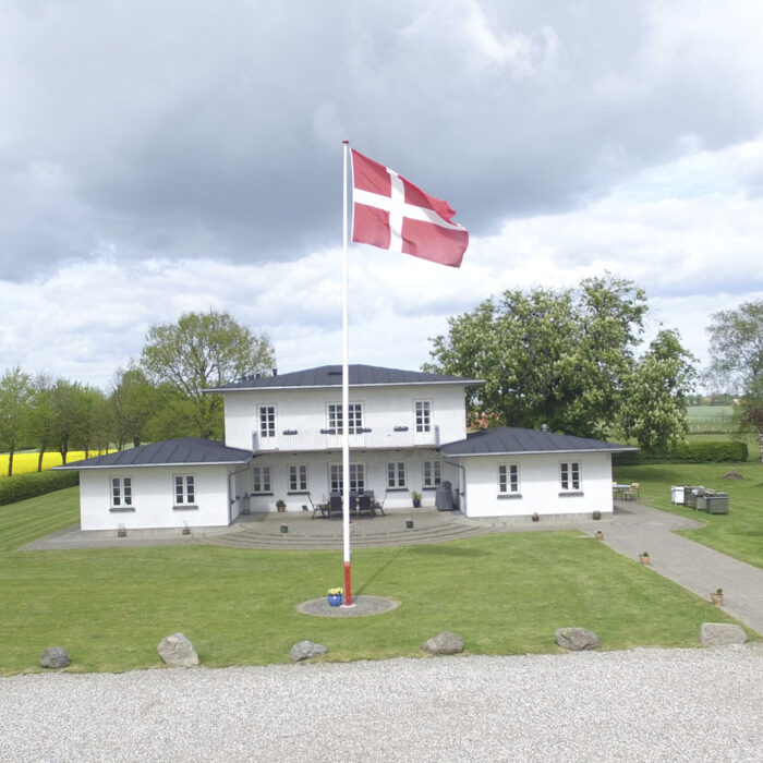 Hvid villa med Danmarks flag i forgrunden. Gule marker på højre side og grønt græs. Vingård.